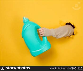 blue plastic bottle with liquid detergent in a female hand on a yellow background. A part of the body sticks out of a torn hole in the background