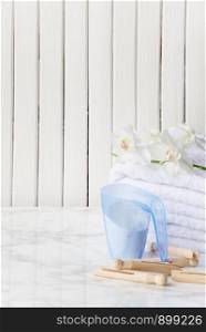 Blue plastic measuring beaker with detergent, stack of white terry towels, white orchid flower and wooden clothespins are on a marble surface against a white background of wooden planks