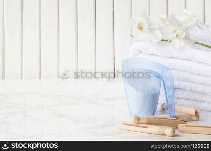 Blue plastic measuring beaker with detergent, stack of white terry towels, wooden clothespins and white orchid flower are on a marble surface against a white background of wooden planks