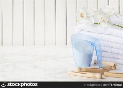 Blue plastic measuring beaker with detergent, stack of white terry towels, wooden clothespins and white orchid flower are on a marble surface against a white background of wooden planks