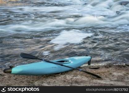 blue plastic white water kayak with a paddle on a river shore with a rapid in background