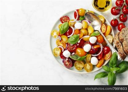 Blue plate of fresh and healthy Mediterranean salad with mozzarella cheese, tomatoes, red onion, basil leaves and a spoon on white marble vintage background.