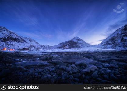 Blue sky with stars and clouds over rocky beach and snow covered mountains at night Lofoten islands, Norway. Beautiful winter landscape with stones, snowy rocks, buildings at dusk. Beautiful Nature