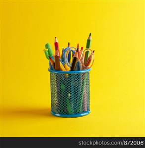 blue stationery glass with multi-colored wooden pencils and pens, yellow background, copy space