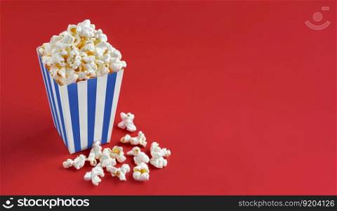 Blue white striped carton bucket with tasty cheese popcorn, isolated on red background. Box with scattering of popcorn grains. Fast food, movies, cinema and entertainment concept.