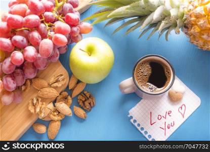 Blue wooden table full with fresh fruits, grapes, pineapple, apple, almonds and walnuts, a cup of coffee and a love you note with heart shaped sugar on it.