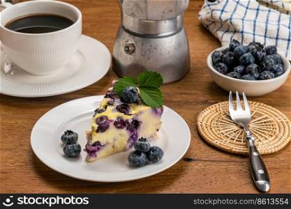Blueberry yogurt cake and black coffee. Slice of homemade blueberry yoghurt cake in white ceramic dish with a cup of black coffee, moka pot coffee maker, blueberries in white ceramic cup and metal fork on bamboo mat on wooden tab≤.