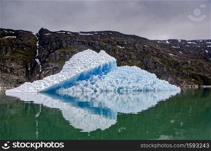 Bluish iceberg in a green sea with mountains in the background