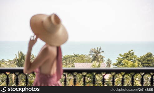 Blur side view of woman in pink dress and straw hat standing on a hotel balcony, ocean view.