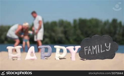 Blurred background of family hugging and kissing on river bank shore. Foreground word happy made of fabric padded letters and word family written with chalk on a black board