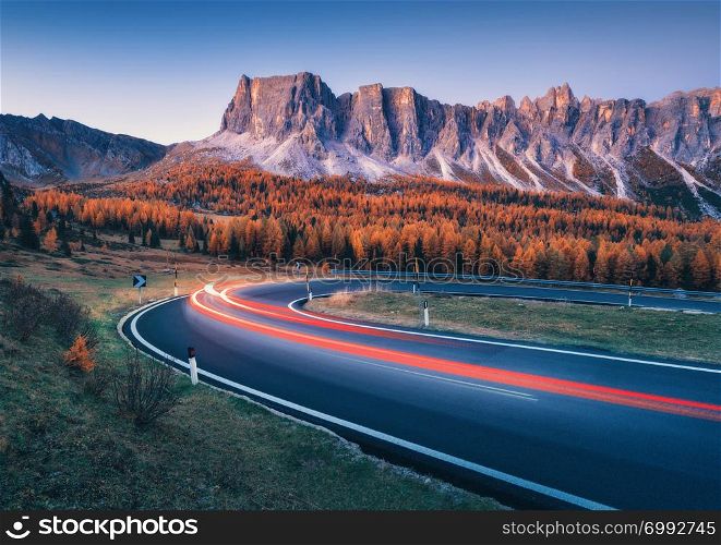 Blurred car headlights on winding asphalt road in mountains at sunset in autumn. Spectacular landscape with light trails, forest, rocks and purple sky at night in fall. Car driving on roadway