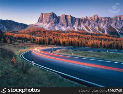 Blurred car headlights on winding asphalt road in mountains at sunset in autumn. Spectacular landscape with light trails, forest, rocks and purple sky at night in fall. Car driving on roadway