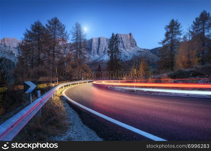 Blurred car headlights on winding road at night in autumn. landscape with asphalt road, light trails, mountains, trees, and blue sky with moon at dusk. Roadway in Italy. View with highway and rocks