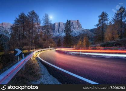 Blurred car headlights on winding road at night in autumn. landscape with asphalt road, light trails, mountains, trees, and blue sky with moon at dusk. Roadway in Italy. View with highway and rocks