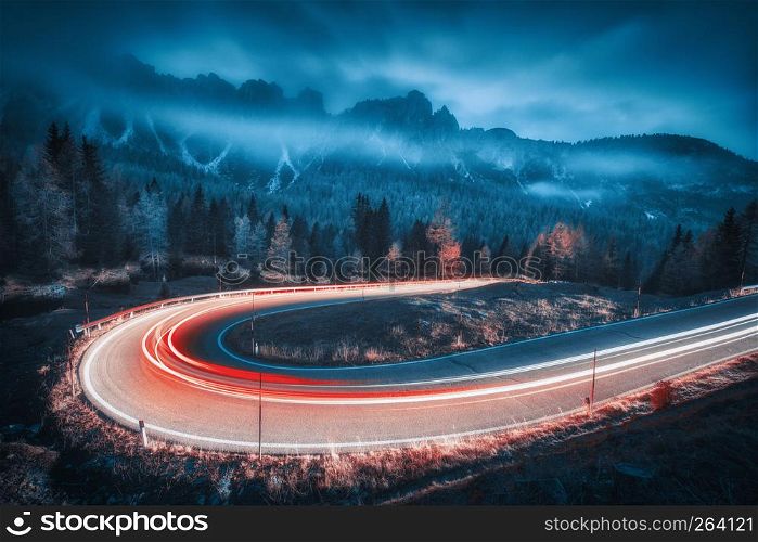 Blurred car headlights on winding road in mountains with low clouds at night in autumn. Moody landscape with asphalt road, light trails, foggy forest, rocks and blue sky at dusk. Roadway in Italy