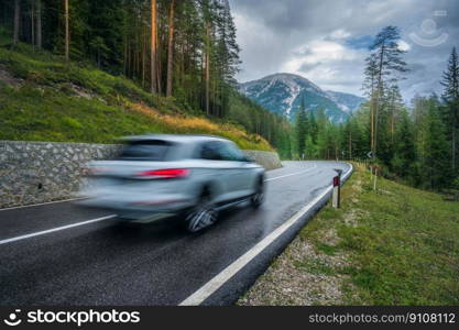 Blurred car in motion on the road in green forest in rain. Dolomites, Italy. Perfect mountain road in overcast rainy summer day. Roadway, pine trees in italian alps. Transportation. Highway in wood