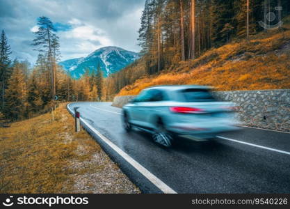 Blurred car in motion on the road in orange forest in rain in autumn. Dolomites, Italy. Perfect mountain road in overcast day in fall. Roadway, pine trees in alps. Transportation. Highway in woods