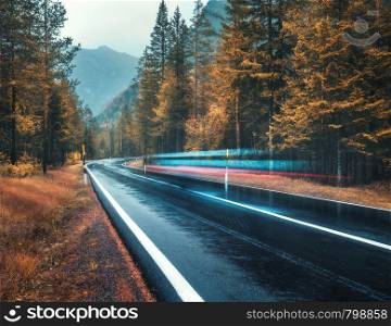 Blurred car on the road in autumn forest in rain. Perfect asphalt mountain road in overcast rainy day. Roadway, orange trees in alps in fall. Nature. Highway in foggy woodland. Car in motion. Travel. Blurred car on the road in autumn forest in rain
