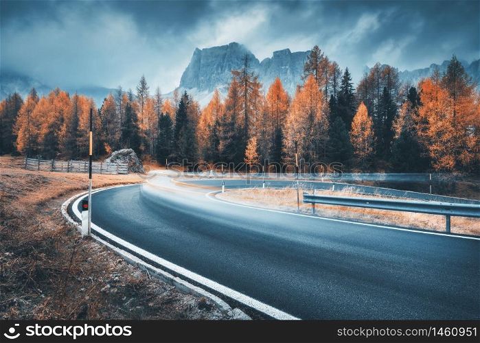 Blurred car on winding road in mountains in overcast rainy evening in autumn. Dramatic landscape with car, orange trees, rocks, blue sky with low clouds at sunset in fall. Car driving on roadway