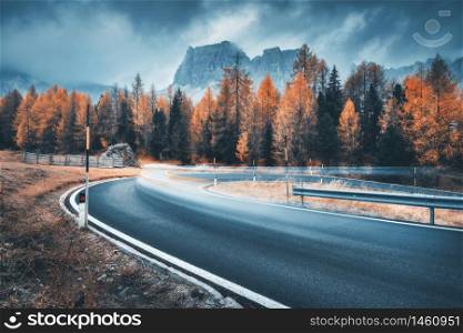 Blurred car on winding road in mountains in overcast rainy evening in autumn. Dramatic landscape with car, orange trees, rocks, blue sky with low clouds at sunset in fall. Car driving on roadway