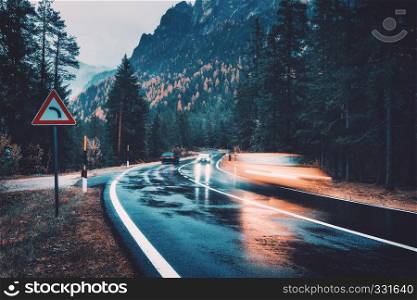 Blurred cars in motion on the road in autumn forest in rain. Perfect asphalt mountain road in overcast rainy day. Roadway, pine trees in italian alps. Transportation. Highway in foggy woodland. Travel
