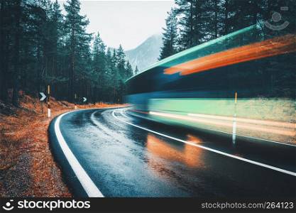 Blurred green bus on the road in autumn forest in rain. Perfect asphalt mountain road in overcast rainy day. Roadway, pine trees in alps. Transportation. Highway in foggy woodland. Car in motion