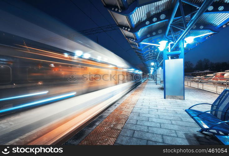 Blurred high speed train on the railway station at night in Europe. Urban landscape with modern passenger train in motion on the railway platform with illumination. Intercity vehicle. Railroad travel