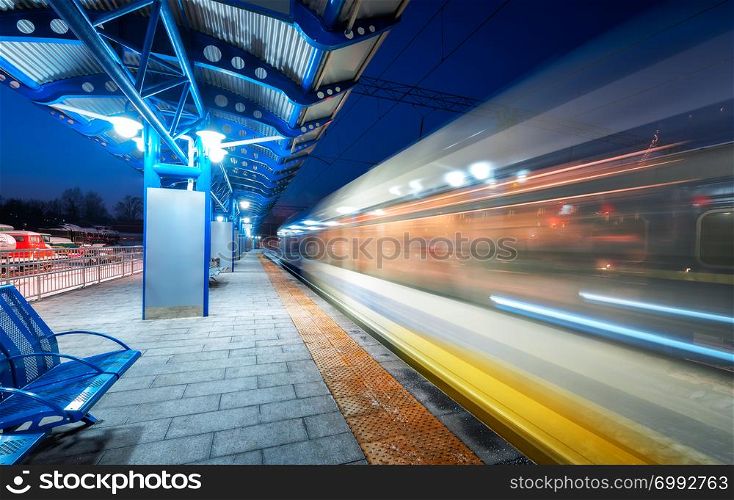 Blurred high speed train on the railway station at night in Europe. Urban landscape with modern passenger train in motion on the railway platform with illumination. Intercity vehicle. Railroad travel