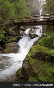 Blurred motion shot of stream and waterfalls in remote wooded area with footbridge