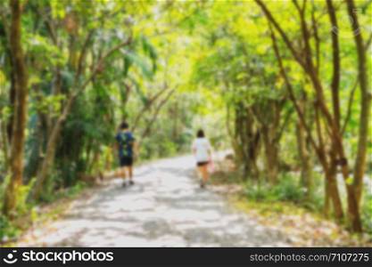 Blurred nature background of two people walking along the pathway in the park, surrounded by lush green trees in summer with natural sunlight.