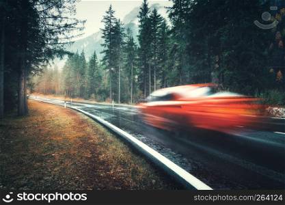 Blurred red car in motion on the road in autumn forest in rain. Perfect asphalt mountain road in overcast rainy day. Roadway, pine trees in italian alps. Transportation. Highway in foggy woodland