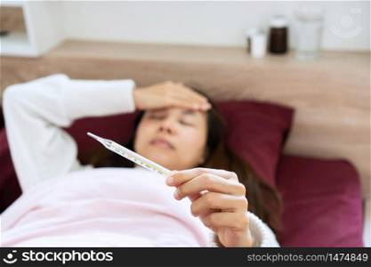 Blurred young woman suffer from fever sick in bed is holding thermometer in hand while using her hand touch at her forehead. he concept of disease and ill