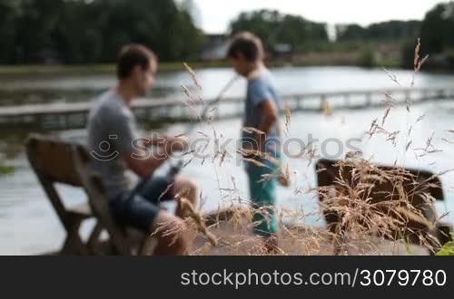 Blurry positive father teaching his boy how to fish while sitting on the chair on wooden pontoon as they spend time together fishing on the lake. Foreground wild plants swaying in the wind. Loving dad and son angling with fishing rods by the pond.