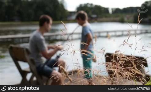 Blurry positive father teaching his boy how to fish while sitting on the chair on wooden pontoon as they spend time together fishing on the lake. Foreground wild plants swaying in the wind. Loving dad and son angling with fishing rods by the pond.
