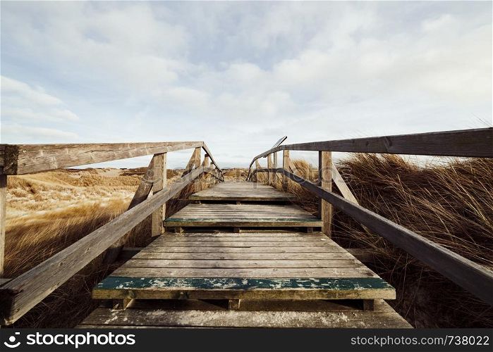 Boardwalk trail crossing seashore landscape with dry grass, viewed from the top of old wooden stairs on the hill. Idyllic picturesque landscape with cloudy sky, Amrum, Germany