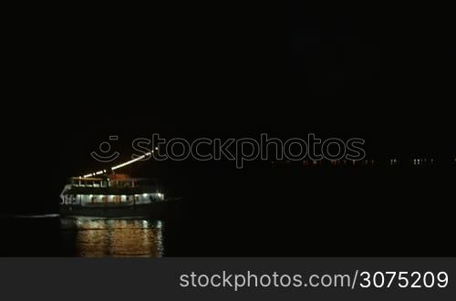 Boat is sailing on the water at night, it is illuminated with lamps. There is a flash of lightning on the sky
