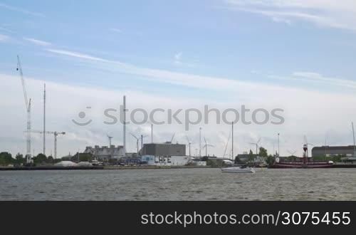 Boat sailing along the shore in the city. Harbour and industrial city district with working windmills