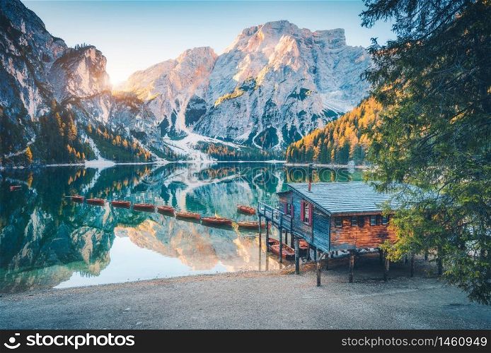 Boats and wooden house on the water on coast of Braies lake at sunrise in autumn in Dolomites, Italy. Landscape with beach, building, mountains, reflection in water, colorful trees in fall. Travel