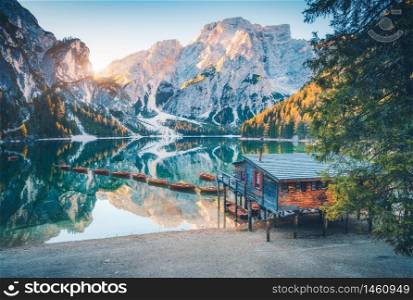 Boats and wooden house on the water on coast of Braies lake at sunrise in autumn in Dolomites, Italy. Landscape with beach, building, mountains, reflection in water, colorful trees in fall. Travel
