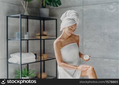 Body care concept. Lovely female model applies moisturising cream on leg, has healthy soft skin after taking bath, wrapped in towel, poses against bathroom interior. Beauty routine, wellness