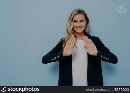 Body language concept. Smiling polite young woman dressed in black blazer over white tshirt keeping hands in stop gesture, saying no or refusing proposal while standing isolated over color background. Smiling polite young woman dressed in black blazer over white tshirt keeping hands in stop gesture
