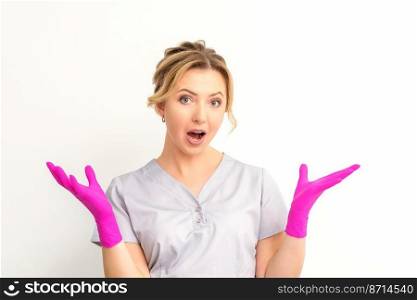 Body language. Young surprised Caucasian woman doctor wearing gloves gesturing with her hands, spread his arms having shocked expression opening her mouth against the white background. Body language. Young surprised Caucasian woman doctor wearing gloves gesturing with her hands, spread his arms having shocked expression opening her mouth against the white background.