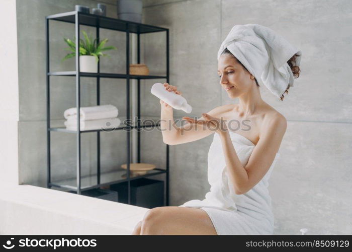 Body lotion applying from bottle. Attractive caucasian girl wrapped in towel after bathing. Happy young woman takes shower at home. Products for smooth silky skin using. Spa procedure.. Body lotion applying from bottle. Happy young woman using products for smooth silky skin.