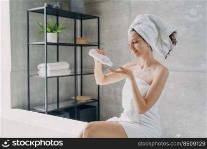 Body lotion applying from bottle. Attractive caucasian girl wrapped in towel after bathing. Happy young woman takes shower at home. Products for smooth silky skin using. Spa procedure.. Body lotion applying from bottle. Happy young woman using products for smooth silky skin.
