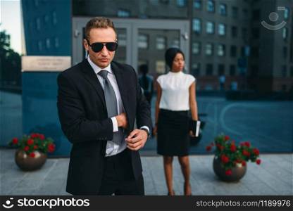 Bodyguard in suit and sunglasses, female VIP client on background. Security guard is a risky profession, professional guarding, business persons protection. Bodyguard in suit and sunglasses, female VIP