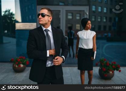 Bodyguard in suit and sunglasses, female VIP client on background. Security guard is a risky profession, professional guarding, business persons protection