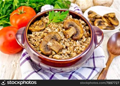 Boiled buckwheat with mushrooms in a brown pottery bowl on a kitchen towel, parsley, tomatoes and fried mushrooms on the background light wooden boards