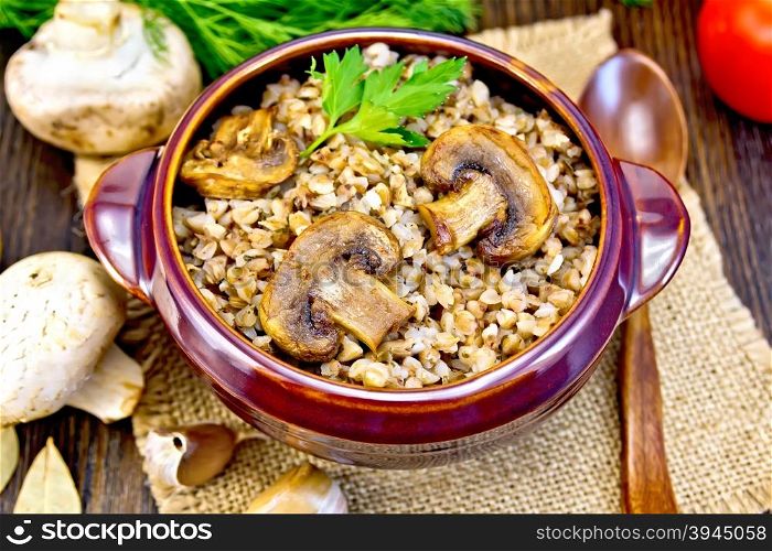 Boiled buckwheat with mushrooms in a brown pottery bowl on a napkin of burlap, parsley, tomatoes and garlic on a dark wooden board