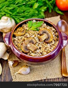 Boiled buckwheat with mushrooms in a brown pottery bowl on a napkin of burlap, parsley, tomatoes and garlic on a wooden boards background