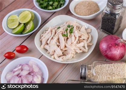 Boiled chicken cut into pieces in a white dish, along with lemons, chilies, lemongrass, red onions, chopped onions, tomatoes, sesame, roasted rice, and pepper on a wooden table.